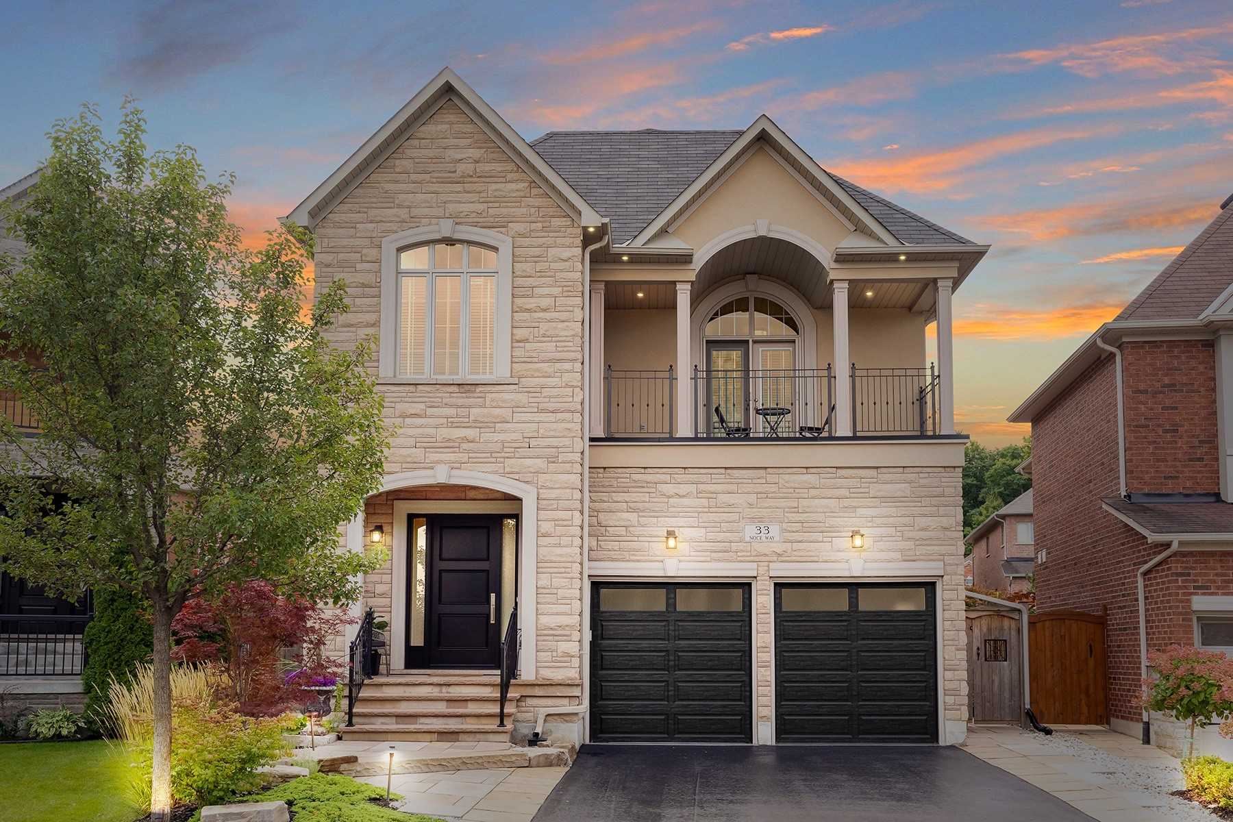 Front exterior of 33 Noce Way at dusk — a stone and stucco detached 2-storey with a black front door, double garage with black doors, and a second-floor balcony with iron railings